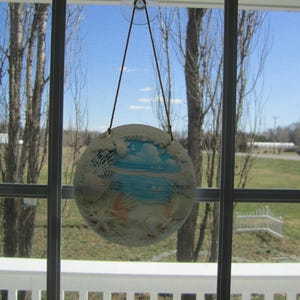 May include: A white, round, decorative hanging ornament with a seashell and beach scene. The ornament is hanging from a window with a suction cup.