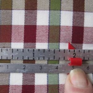 May include: A metal ruler with metric measurements is placed over a red, white, brown, and green plaid fabric. A small red plastic piece is positioned near the 5-inch mark on the ruler. The fabric has a classic checkered pattern.