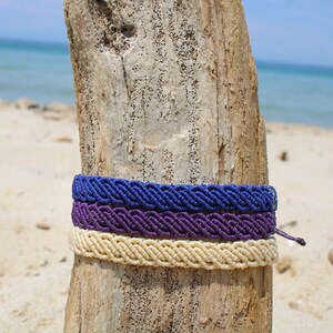 May include: Three braided bracelets in shades of purple and cream are tied around a weathered piece of driftwood. The driftwood is on a sandy beach with the ocean in the background.