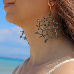 May include: A close-up of a person wearing a pair of handmade, macrame earrings. The earrings are a dark green colour with gold-coloured beads and gold-coloured ear hooks. The earrings are in a snowflake design.