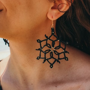 May include: Black snowflake earrings with gold-coloured detailing. The earrings are crafted from a woven material and suspended from gold-coloured hooks. A distinctive accessory.