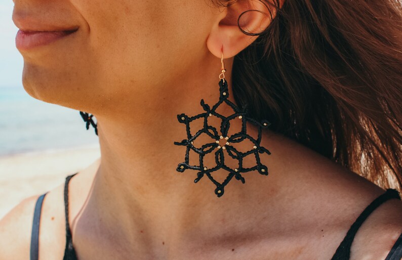 May include: Black snowflake earrings with gold-coloured detailing. The earrings are crafted from a woven material and suspended from gold-coloured hooks. A distinctive accessory.
