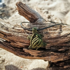 Puede incluir: Un colgante de tortuga de cuerda anudada verde y negra en un cordón negro, descansando sobre un trozo de madera a la deriva.
