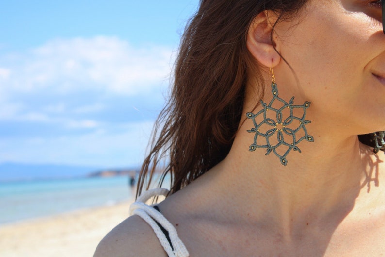 May include: A close-up of a snowflake-shaped earring. The earring is a dark teal colour with gold-coloured accents. The earring is hanging from a gold-coloured hook. The background is a beach with blue sky and water.