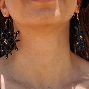 May include: Black macram&eacute; earrings with gold-coloured accents. The earrings feature a geometric design with a circular shape and a dangling rectangular shape. They are suspended from gold-coloured hooks.