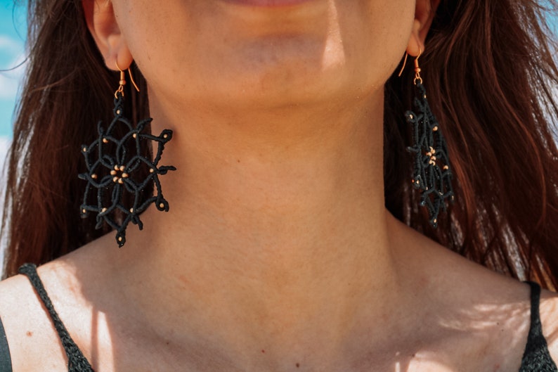 May include: Black macram&eacute; earrings with gold-coloured accents. The earrings feature a geometric design with a circular shape and a dangling rectangular shape. They are suspended from gold-coloured hooks.