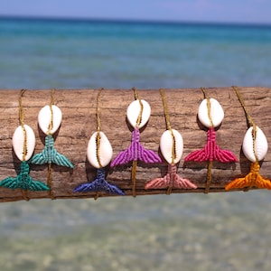 May include: Eight colorful macrame mermaid tail bracelets with white cowrie shells. The bracelets are displayed on a piece of driftwood against a blue ocean background.