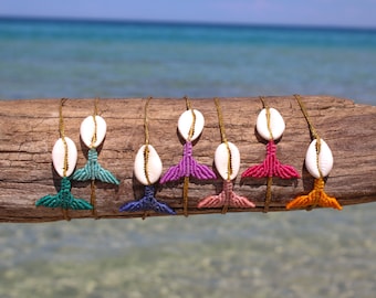 May include: Eight colourful macrame mermaid tail bracelets with white cowrie shells. The bracelets are displayed on a piece of driftwood against a blue ocean background.