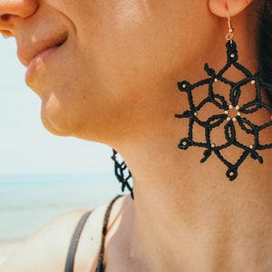 May include: Black macram&eacute; earrings in a snowflake design. The earrings have a geometric pattern with small gold beads. They are suspended from gold-coloured ear wires. The background is a blurred view of the sea and sky.
