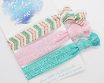 May include: A set of three hair ties in pastel colors: pink, teal, and a patterned chevron design with gold accents. The ties are displayed on a white card with the text "White Wave Boutique".