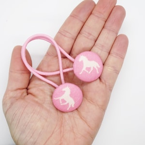 May include: Two pink hair ties with white horse silhouettes. The hair ties are made of fabric and have a button-like design.