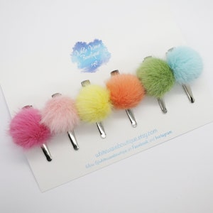 May include: Six colourful faux fur pom pom hair clips. The clips are arranged in a rainbow order from pink to light blue.  The clips are on a white background with the text "White Wave Boutique" and "follow @whitewaveboutique on Facebook and Instagram".