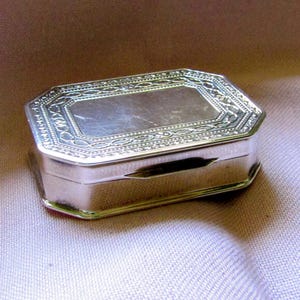 Puede incluir: Una caja rectangular de metal plateado con esquinas biseladas y tapa con bisagras. La superficie superior presenta un intrincado patrón de borde grabado que rodea un panel central liso y sencillo. Los lados de la caja son lisos y pulidos.
