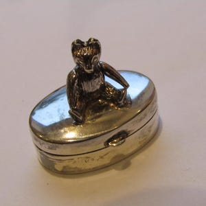 Könnte beinhalten: Kleine, silberfarbene Pillendose mit Klappdeckel. Auf der Oberseite ist eine detaillierte, sitzende Bärenfigur angebracht. Die Dose hat eine ovale Form und eine glänzende, reflektierende Oberfläche. Der Bär hat eine dunkle, metallische Farbe.