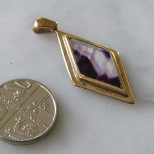 Puede incluir: Un colgante de oro con una piedra de color púrpura y blanco en forma de diamante.