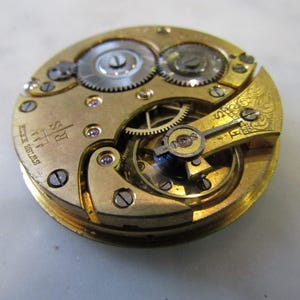 May include: Close-up of a gold-coloured pocket watch movement with intricate gears and an 'R.S. Swiss 17 Jewels' inscription.