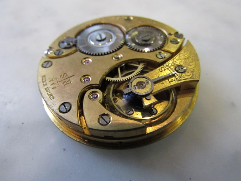 May include: Close-up of a gold-coloured pocket watch movement with intricate gears and an 'R.S. Swiss 17 Jewels' inscription.
