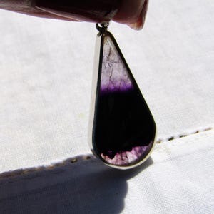 Puede incluir: Colgante en forma de lágrima con marco plateado. La piedra pulida presenta un degradado de color, pasando del morado oscuro en la parte inferior al morado claro y blanco en la parte superior. El colgante está suspendido de un pequeño lazo plateado.