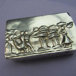 May include: A silver rectangular box with an embossed design of people dancing around a tree. The box has a textured surface and a raised edge.