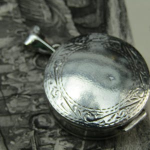 May include: A silver-coloured, round locket with an ornate, engraved floral design. The locket has a small loop at the top for a chain and a clasp on the side. The background is a vintage print.