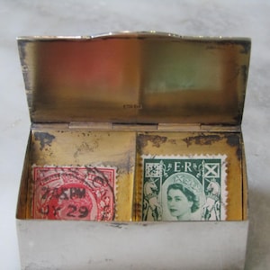 Könnte beinhalten: Eine silberne Schachtel mit Scharnieren und zwei Fächern, die jeweils einen alten Briefmarken enthalten. Die linke Marke ist rot und trägt die Nummer "29". Die rechte Marke ist grün und zeigt das Porträt einer Frau mit dem Text "E.R." über ihrem Kopf.