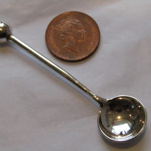 May include: A small, silver-coloured spoon with a rounded bowl and a ball-shaped end. A copper coin is next to the spoon. The spoon is likely used for measuring small amounts of ingredients.