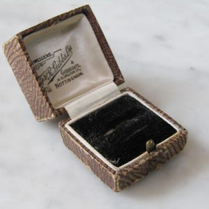 Puede incluir: Una caja de anillo marrón con un interior de terciopelo negro. La caja está abierta y la tapa tiene el texto "Jewellers, J.R. Gibbs & Co., 4 Goosegate, Nottingham" impreso en ella.