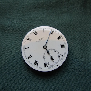 K&ouml;nnte beinhalten: Ein Vintage-Taschenuhrzifferblatt mit wei&szlig;em Hintergrund, schwarzen r&ouml;mischen Ziffern und einem kleineren Sekundenzeiger. Die Zeiger der Uhr sind schwarz und blau. Der Text "J.W. Benson London" ist auf dem Zifferblatt aufgedruckt.