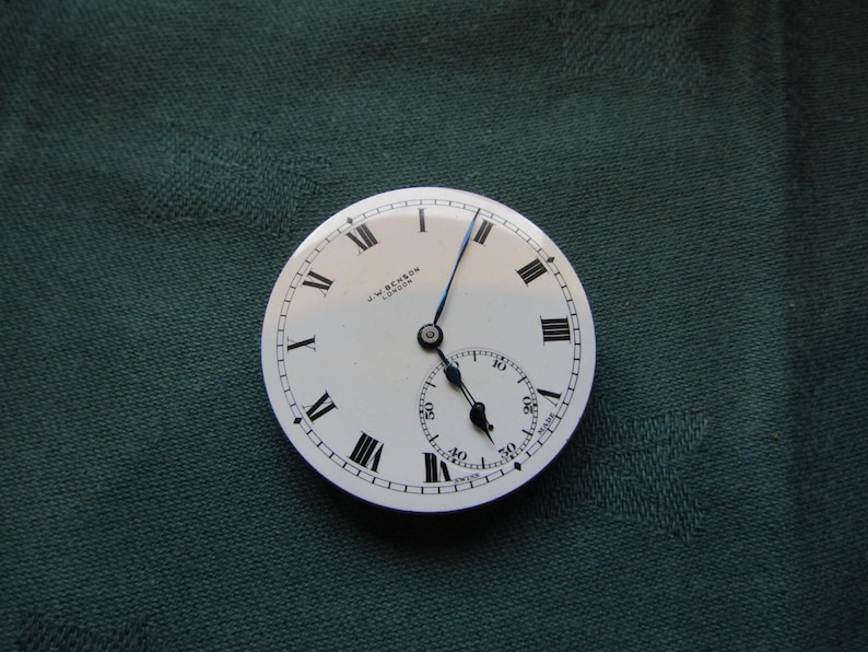 K&ouml;nnte beinhalten: Ein Vintage-Taschenuhrzifferblatt mit wei&szlig;em Hintergrund, schwarzen r&ouml;mischen Ziffern und einem kleineren Sekundenzeiger. Die Zeiger der Uhr sind schwarz und blau. Der Text "J.W. Benson London" ist auf dem Zifferblatt aufgedruckt.