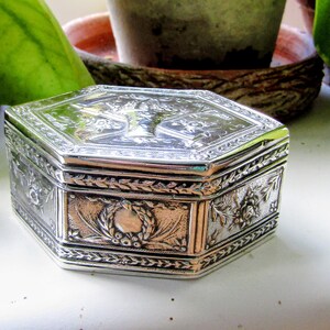 Puede incluir: Un joyero hexagonal de color plateado con intrincados patrones florales en relieve y un motivo central de cesta en la tapa. Los lados de la caja están decorados con diseños detallados de coronas de laurel y bordes de hojas en metal brillante.