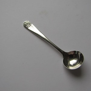 Puede incluir: Una pequeña cuchara plateada con un cuenco redondeado y un diseño decorativo de concha en el mango. La cuchara probablemente está hecha de plata de ley o un metal similar. Diseñada para servir sal u otras porciones pequeñas.