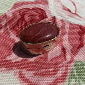 May include: A small, oval-shaped trinket box with a red, marbled lid. The box is made of metal and has a silver finish.
