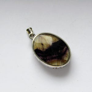 Puede incluir: Colgante ovalado con engaste plateado. El colgante presenta una piedra pulida con una mezcla de colores, incluyendo crema, marrón y morado oscuro. El colgante tiene un pequeño lazo en la parte superior para una cadena.