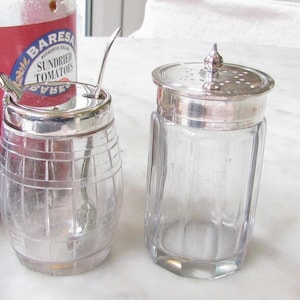 May include: Two glass jars with silver lids. One is barrel-shaped with a spoon inside, the other is rectangular with a perforated lid. A jar of sundried tomatoes is partially visible in the background.