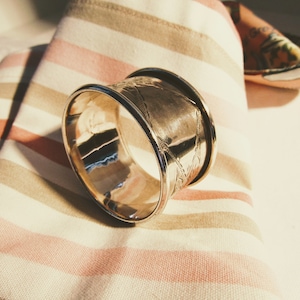 May include: Silver napkin ring with an ornate design. The ring is on a striped fabric background.