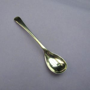 May include: A silver teaspoon with a rounded bowl. The spoon is on a light purple background.
