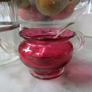 May include: A small, ruby red glass creamer with a handle and a silver teaspoon inside. The creamer is sitting on a white surface.