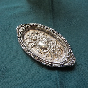 May include: An oval, silver-colored dish with ornate floral and scrollwork detailing. The dish has a raised, decorative border and a textured interior. The background is a dark teal fabric.