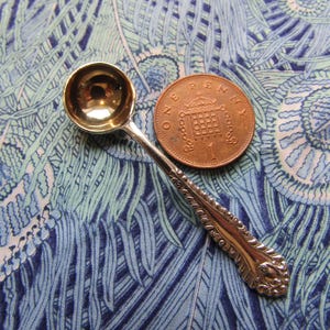 Puede incluir: Una pequeña cuchara plateada ornamentada, con un cuenco redondo y un mango decorativo. Una moneda de cobre de 1 penique está al lado de la cuchara. La cuchara y la moneda descansan sobre una tela estampada azul y verde.