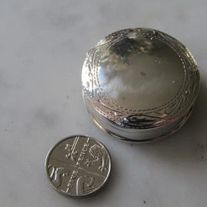 May include: A silver round trinket box with an ornate design. The box is about the size of a UK 20 pence coin.