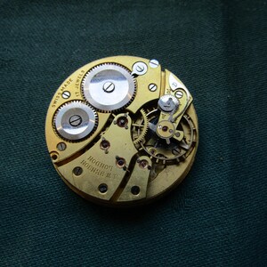 K&ouml;nnte beinhalten: Nahaufnahme eines Vintage-Uhrwerks mit komplizierten Zahnr&auml;dern und Komponenten. Der goldfarbene Mechanismus tr&auml;gt die Aufschrift "SWISS MADE 17 JEWELS" und "A.W. BENSON LONDON". Das Uhrwerk liegt auf einem dunkelgr&uuml;nen Hintergrund.