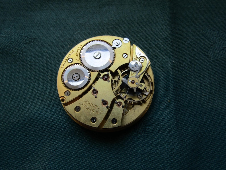 K&ouml;nnte beinhalten: Nahaufnahme eines Vintage-Uhrwerks mit komplizierten Zahnr&auml;dern und Komponenten. Der goldfarbene Mechanismus tr&auml;gt die Aufschrift "SWISS MADE 17 JEWELS" und "A.W. BENSON LONDON". Das Uhrwerk liegt auf einem dunkelgr&uuml;nen Hintergrund.