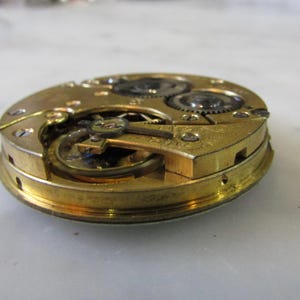 May include: Close-up of a gold-toned pocket watch movement with intricate gears and a winding mechanism.