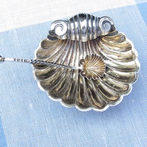 May include: A silver seashell-shaped dish with a decorative spoon. The dish has a scalloped edge and a gold-coloured shell accent in the centre. The spoon has a twisted handle and a small, round end.