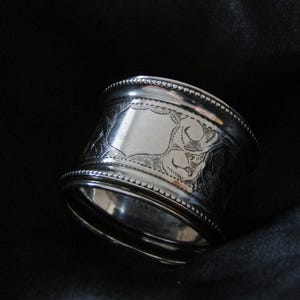 May include: A silver napkin ring with an engraved floral design. The ring has a wide band with a beaded edge and a rectangular cartouche. The background is dark.
