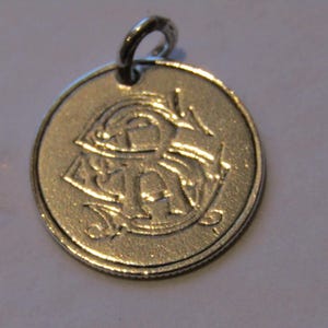 Puede incluir: Un colgante redondo de color plateado con un lazo en la parte superior. El colgante presenta un diseño en relieve de letras y formas estilizadas. El diseño general es simple y elegante, adecuado para joyería o proyectos de artesanía.