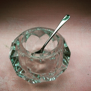 May include: A clear glass salt cellar with a silver spoon inside. The cellar has a faceted design and is sitting on a pink surface.