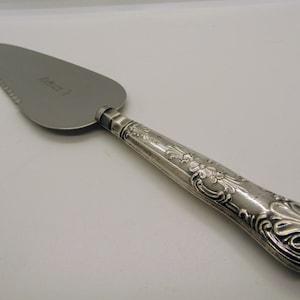 May include: A silver cake server with a serrated blade and ornate handle. The handle features floral and scroll designs. The blade is approximately 23 cm long. The server is likely made of stainless steel or a similar metal.