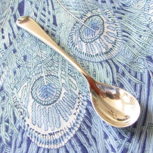 May include: A polished silver-toned metal spoon with a smooth, rounded handle and a deep, oval-shaped bowl. The spoon features a reflective, metallic surface that catches light, showcasing a simple, clean design with no decorative engravings.
