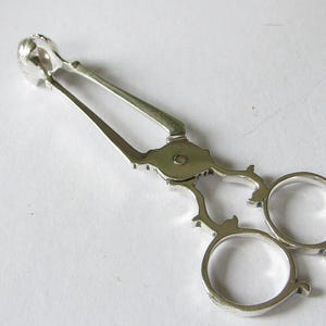 Puede incluir: Pinzas de azúcar plateadas con un diseño decorativo. Las pinzas tienen un mecanismo de tijera con aros para los dedos y un extremo en forma de garra para agarrar los terrones de azúcar. Diseño ornamentado y vintage.